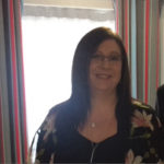 a lady stood by the window with dark hair and flowers on her top